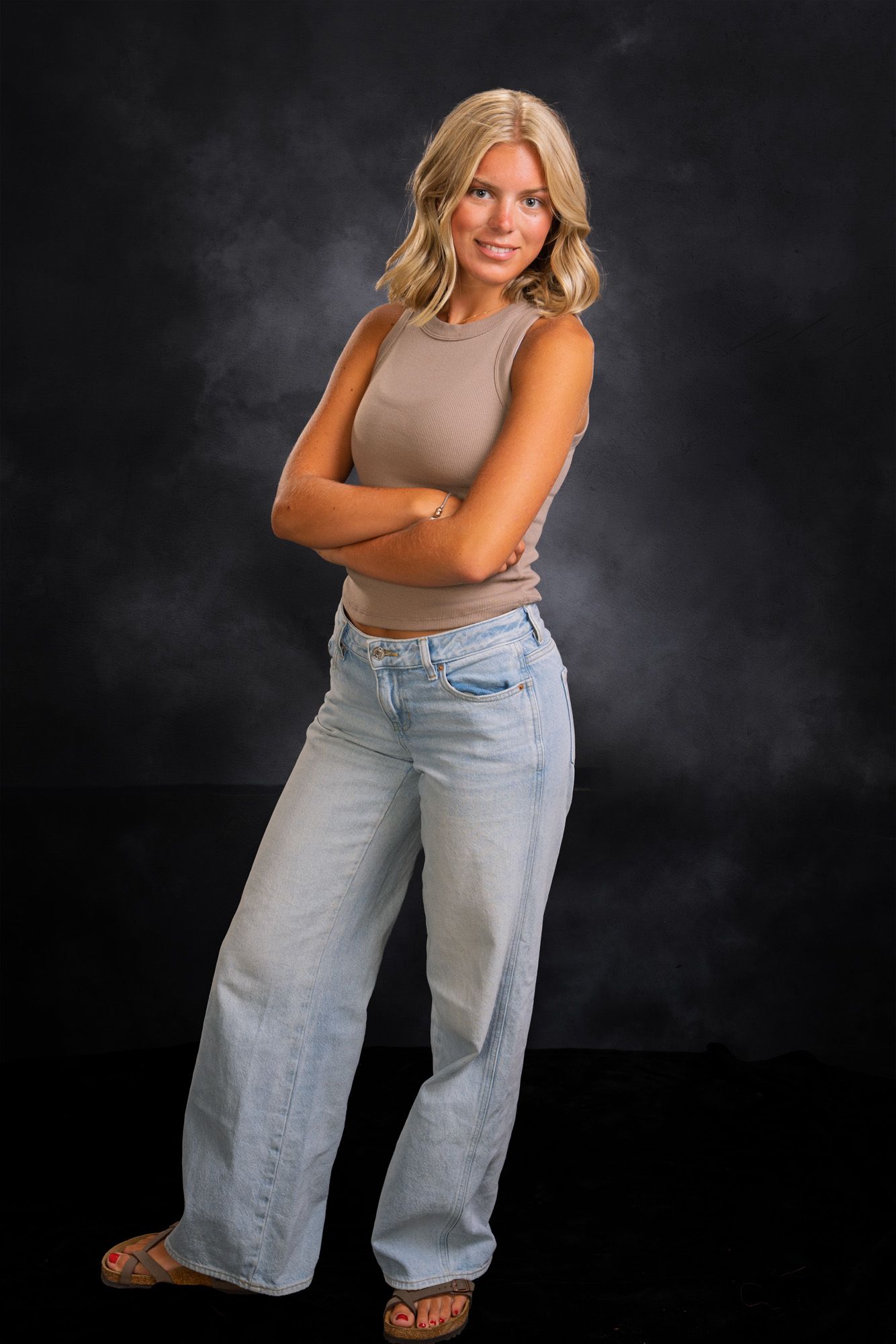 A confident blonde woman poses with arms crossed wearing a tan tank top and wide-leg jeans against a dark backdrop.