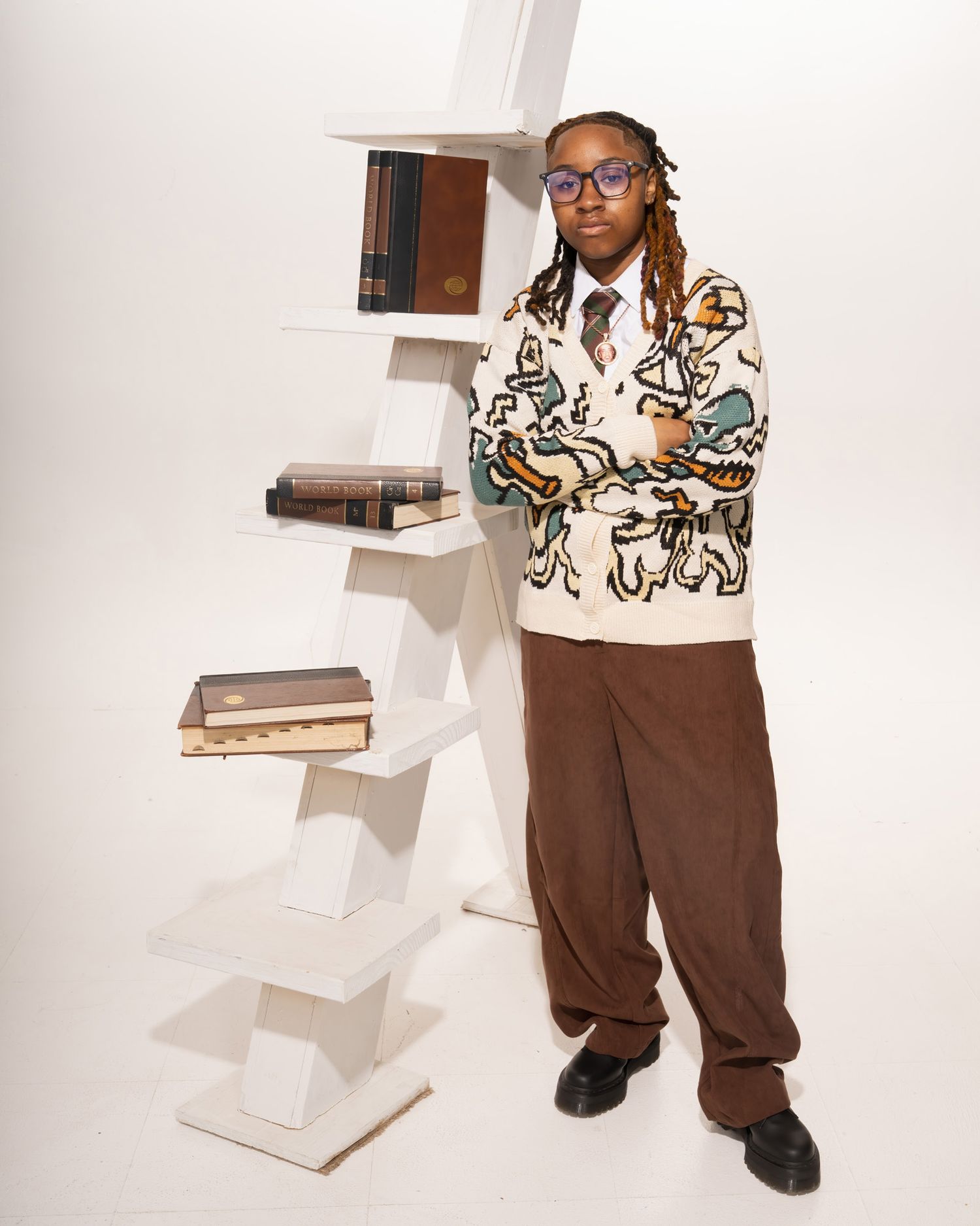 Student standing beside white geometric bookshelf in patterned cardigan and brown trousers, studio portrait.