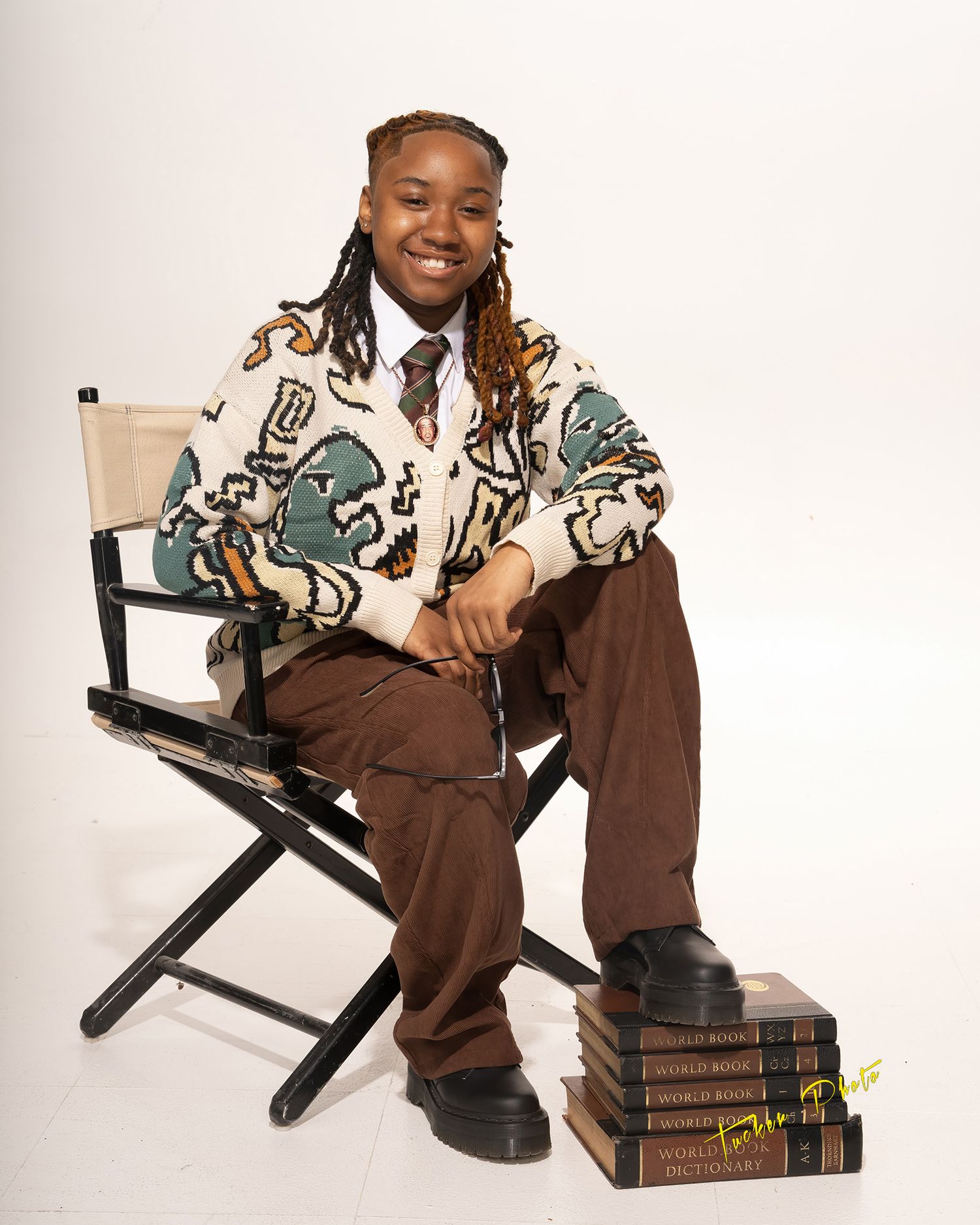Young person sitting in a directors chair wearing a patterned cardigan with stacked books.