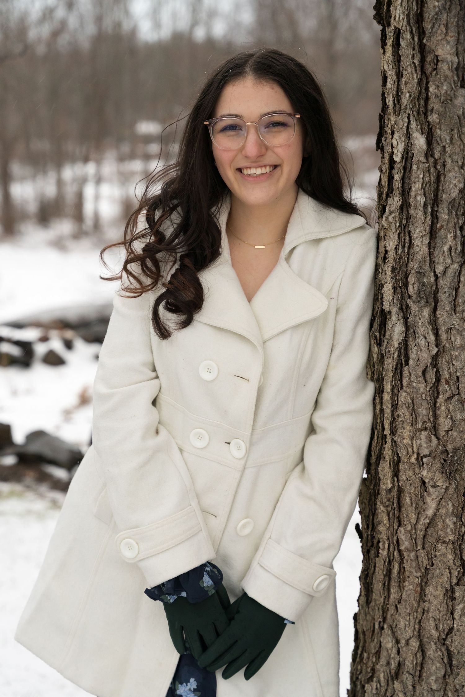 Smiling young woman in a white coat and green gloves posing by a tree in winter snow.