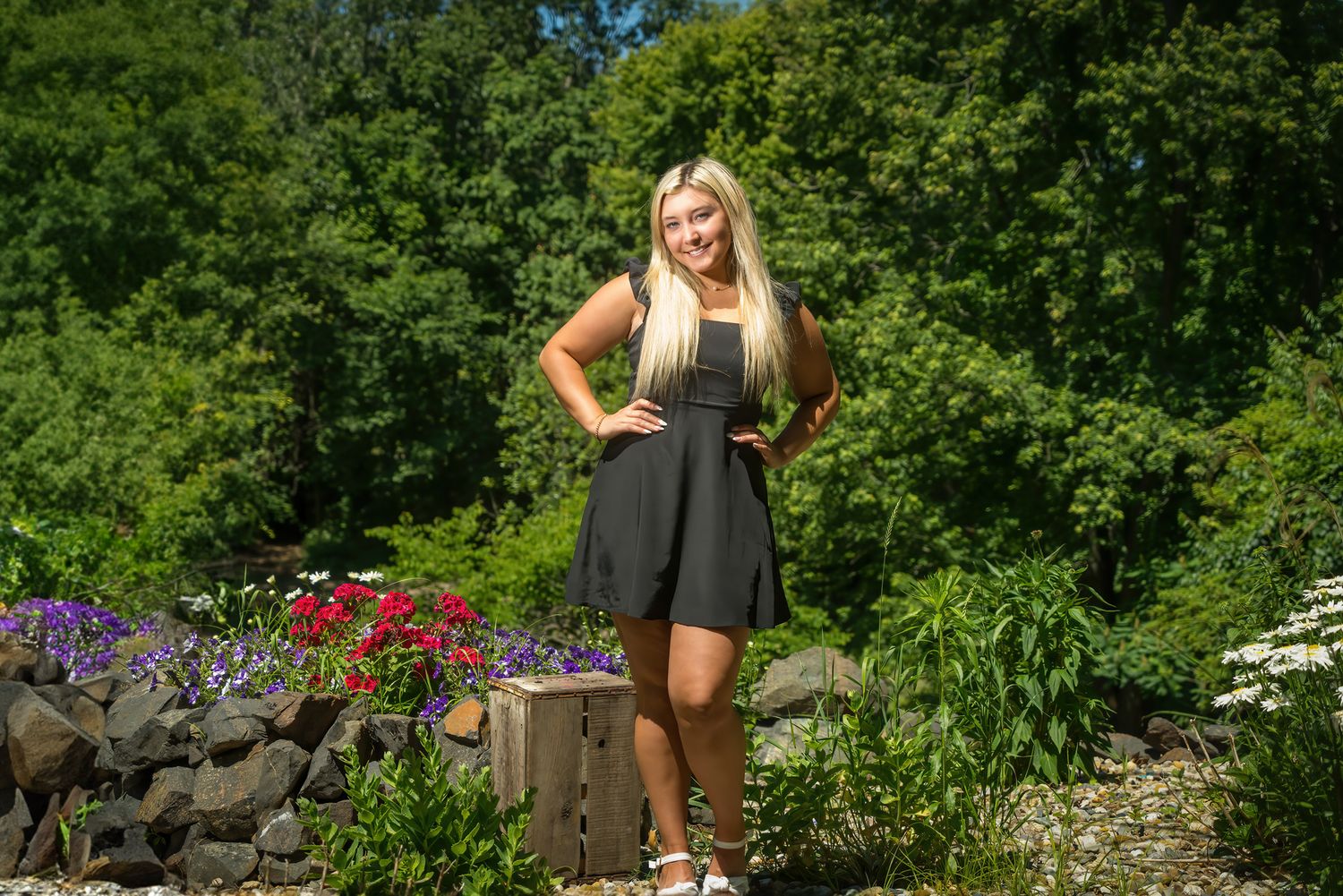 Blonde teenage girl in black dress posing in front of colorful garden for senior portraits.