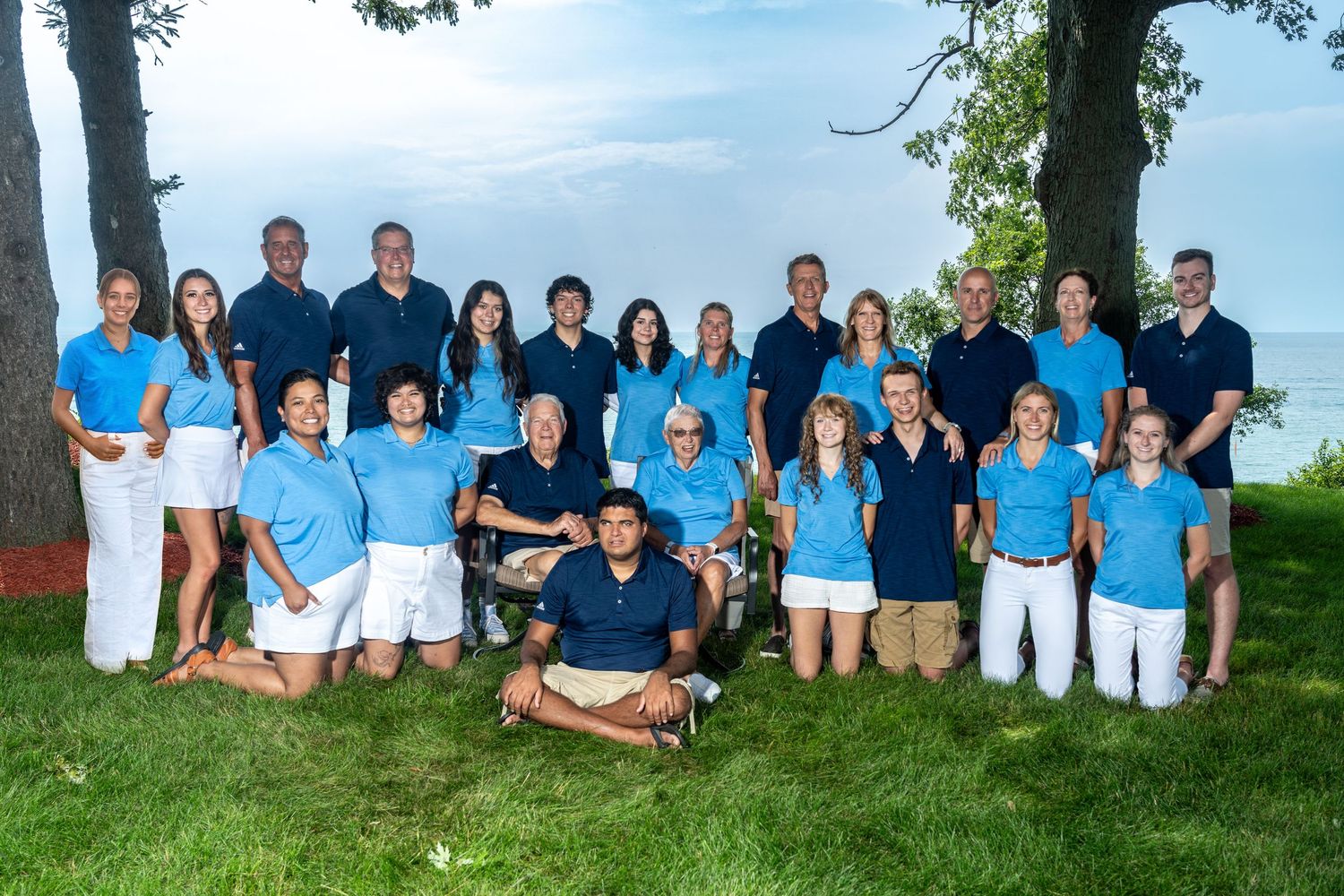 Large group of adults in matching blue polo shirts posing outdoors near lake for team photo.