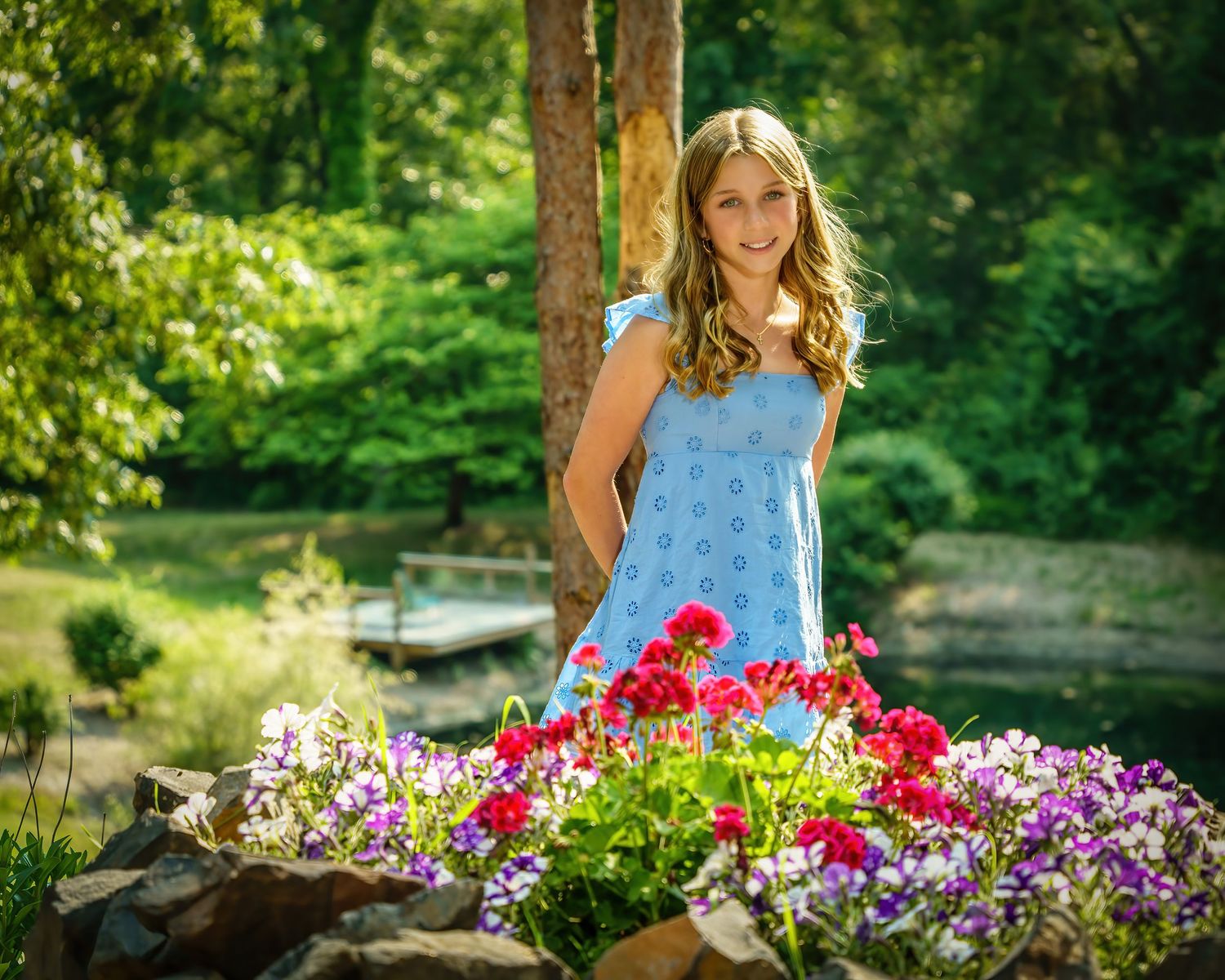 Smiling preteen girl in blue dress standing near colorful garden flowers by a pond.