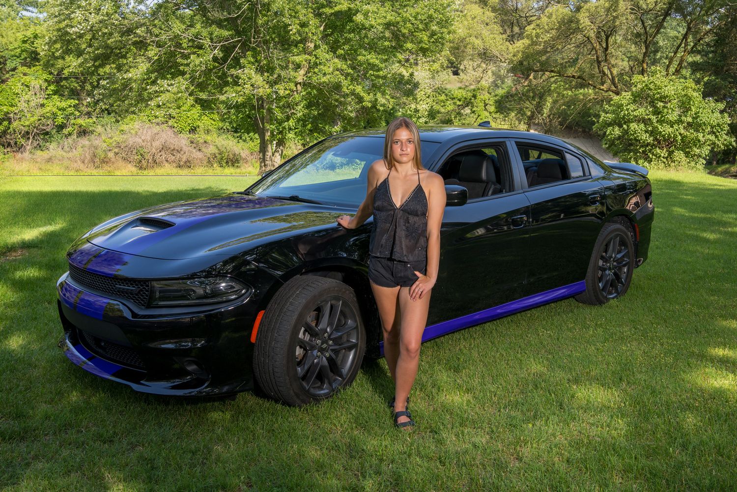 Young woman posing beside a black Dodge Charger with blue accents on green grass.