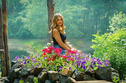 Blonde teenage girl posing among colorful flowers in a misty woodland garden setting.