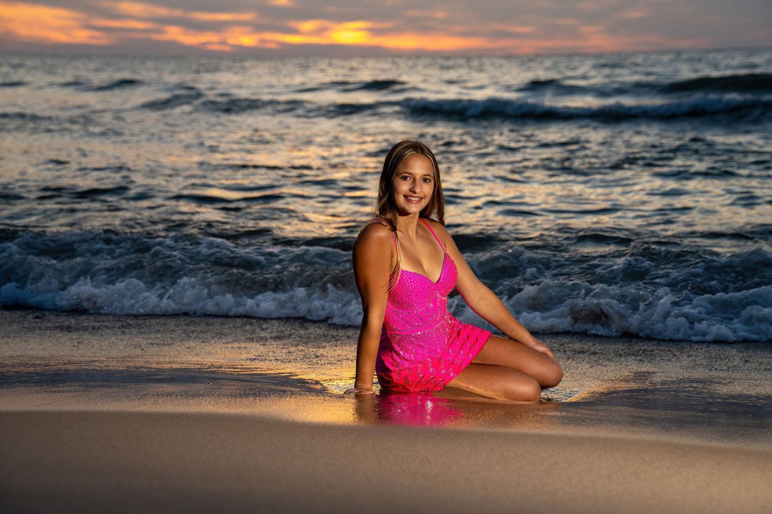 Teen girl in bright pink dress sitting on beach shoreline with crashing waves at sunset.