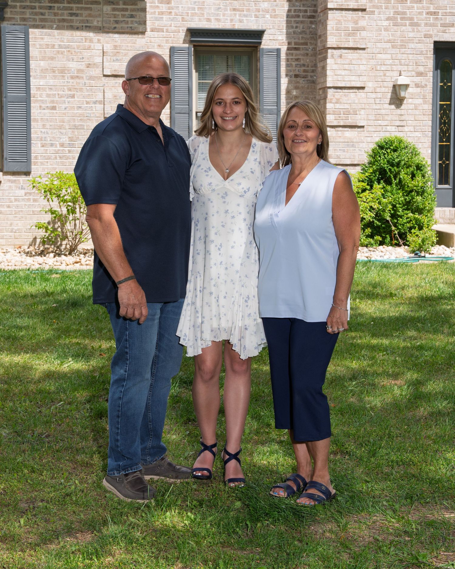 A teenage girl in a floral dress poses with her parents on a sunny lawn in front of a brick house.