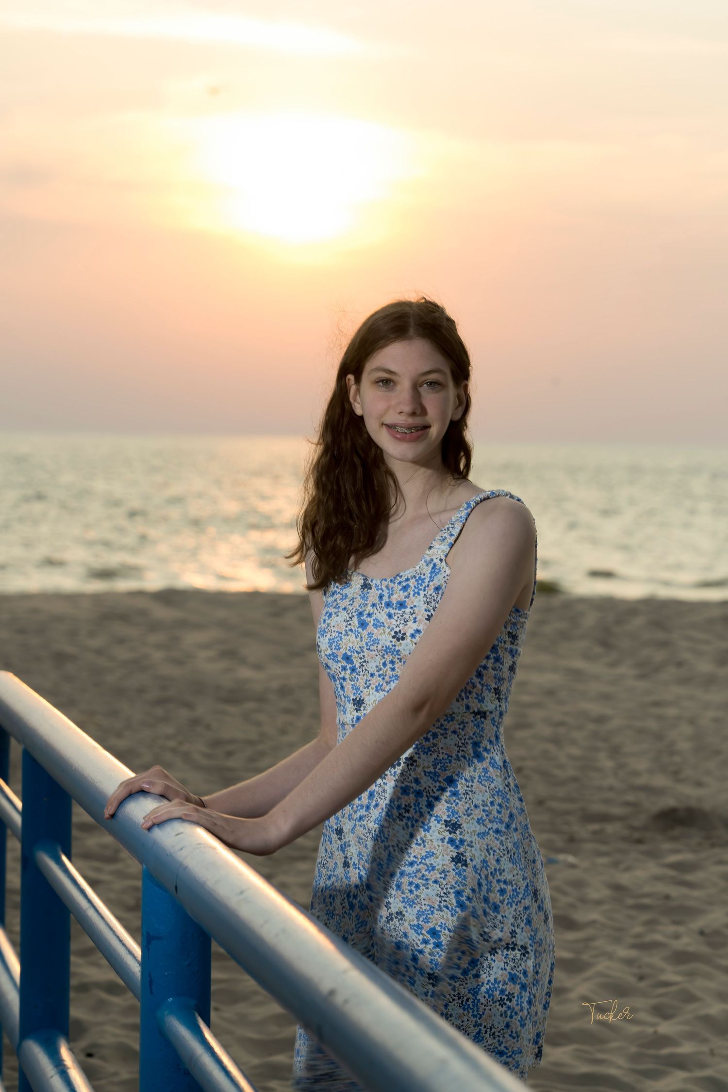 Young woman in blue floral sundress standing at beach railing with golden sunset and ocean in background.