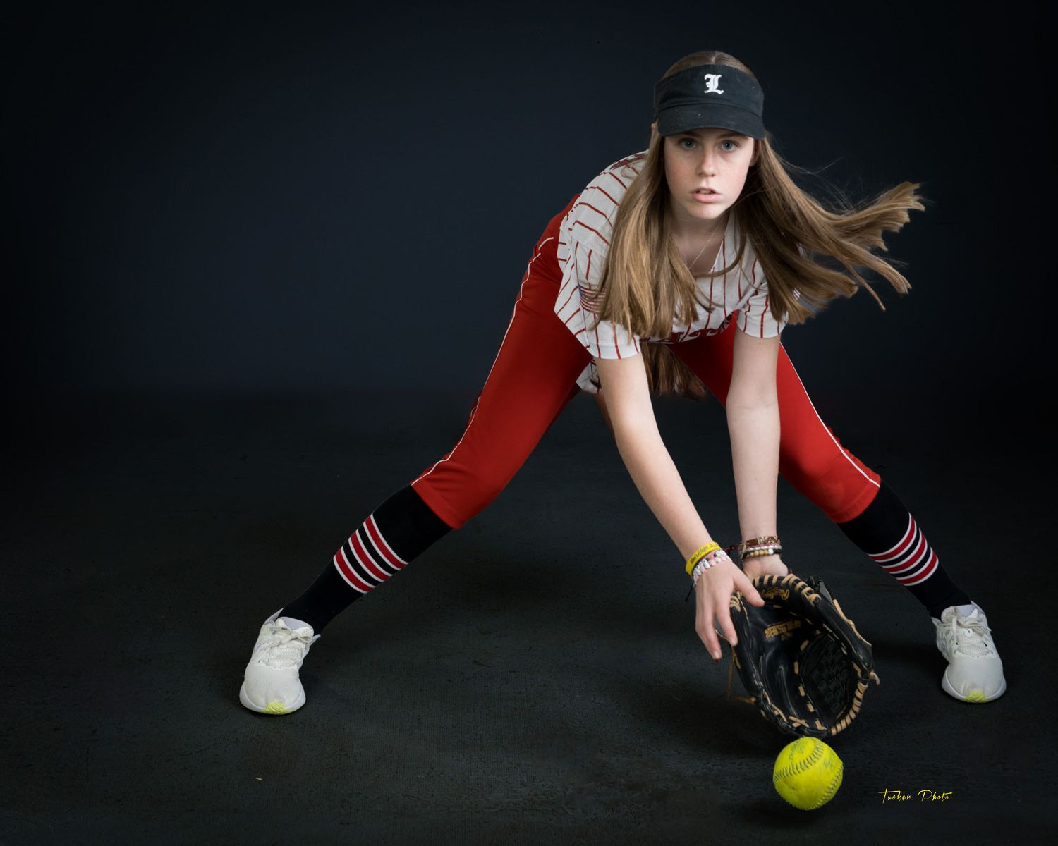Teen softball player fielding a yellow ball in red uniform on dark background.