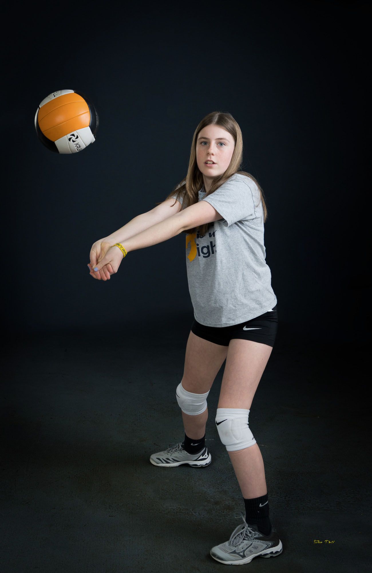Teen girl in volleyball stance bumping an orange and black ball wearing gray shirt and knee pads.