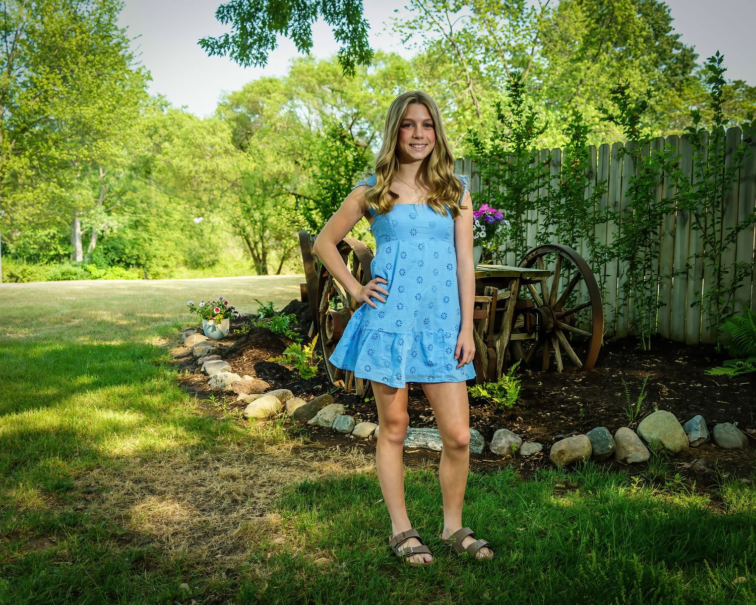 Young woman in blue floral sundress posing in lush green backyard garden with vintage wagon wheel decor.