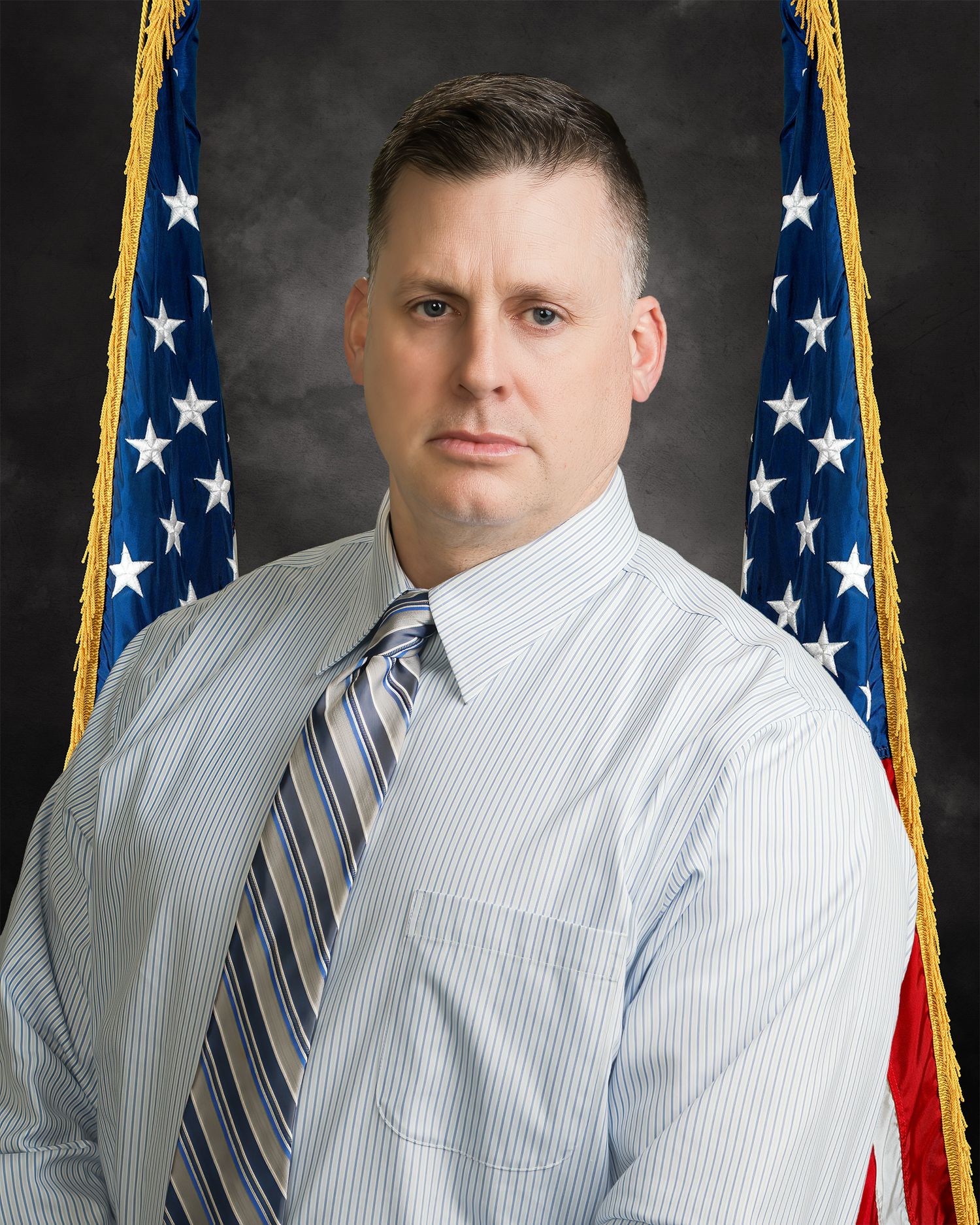 Professional law enforcement official in striped dress shirt and tie posing before an American flag backdrop.