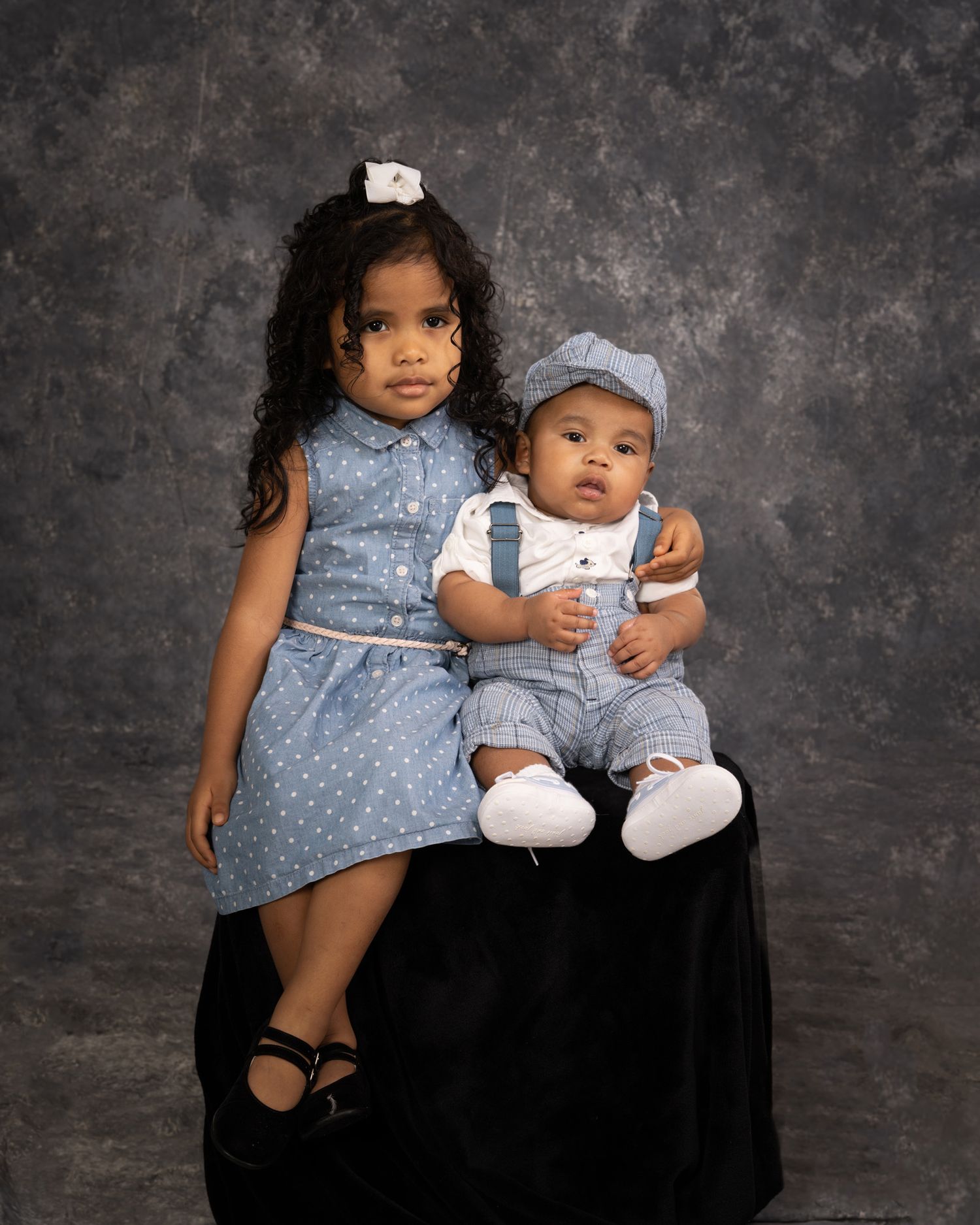 Young girl in blue polka dot dress posing with baby brother in studio portrait.