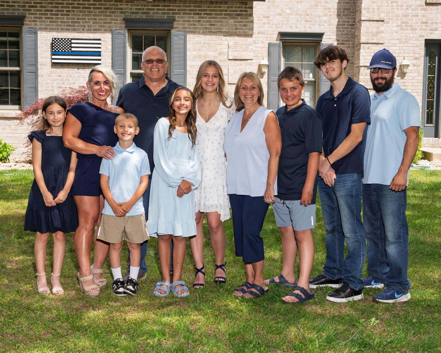A multigenerational family group photo taken outdoors on a green lawn in front of a brick house.