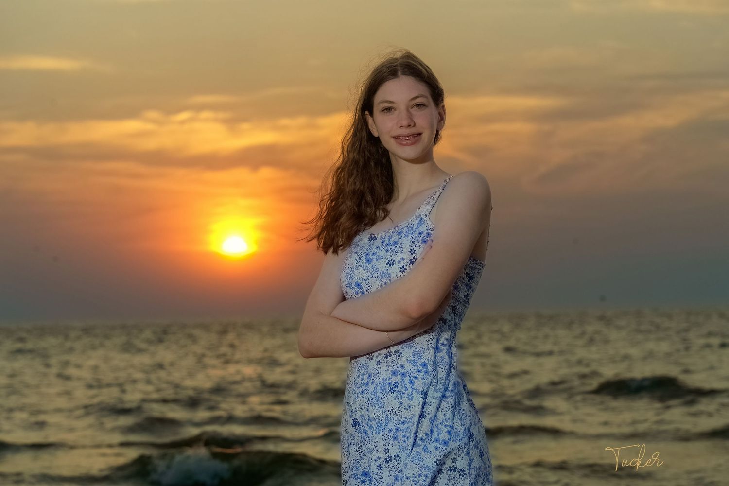 Young woman in blue floral dress posing with arms crossed at beach during golden sunset.
