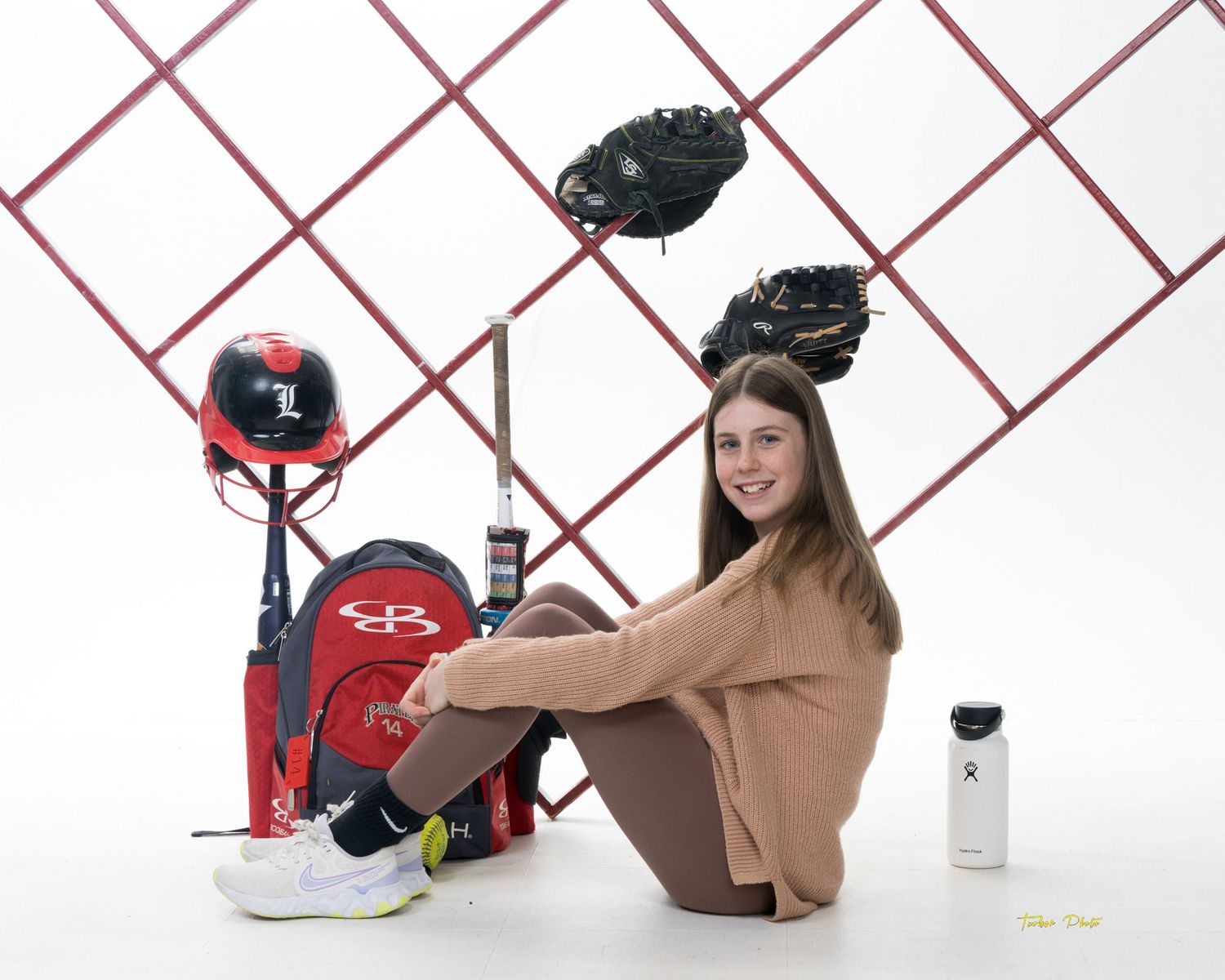 Young female softball player sits beside her Pirates team gear including backpack, helmet, and bat in a studio setting.