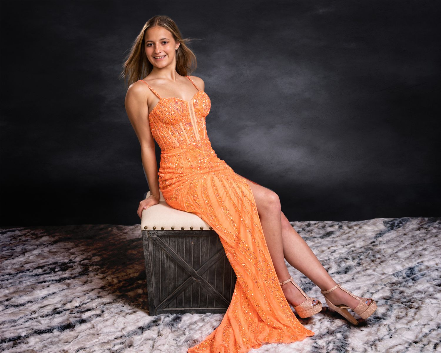 Teen girl in orange lace gown sits gracefully on wooden crate.