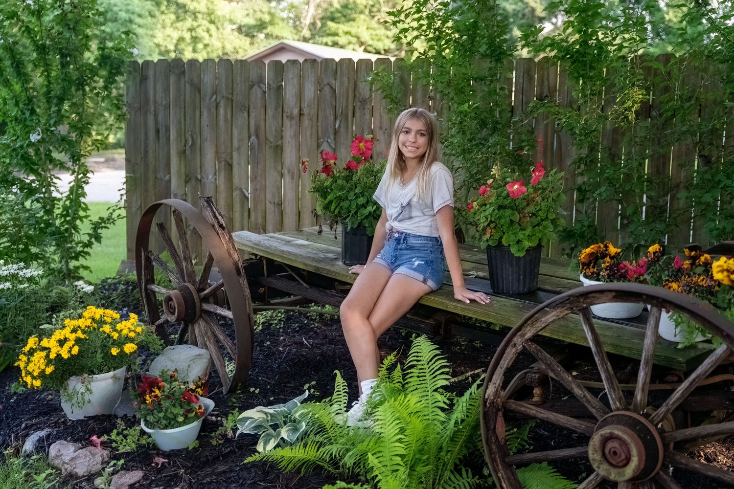 Blonde teen girl in white tee and denim shorts poses on rustic wooden wagon in garden setting.
