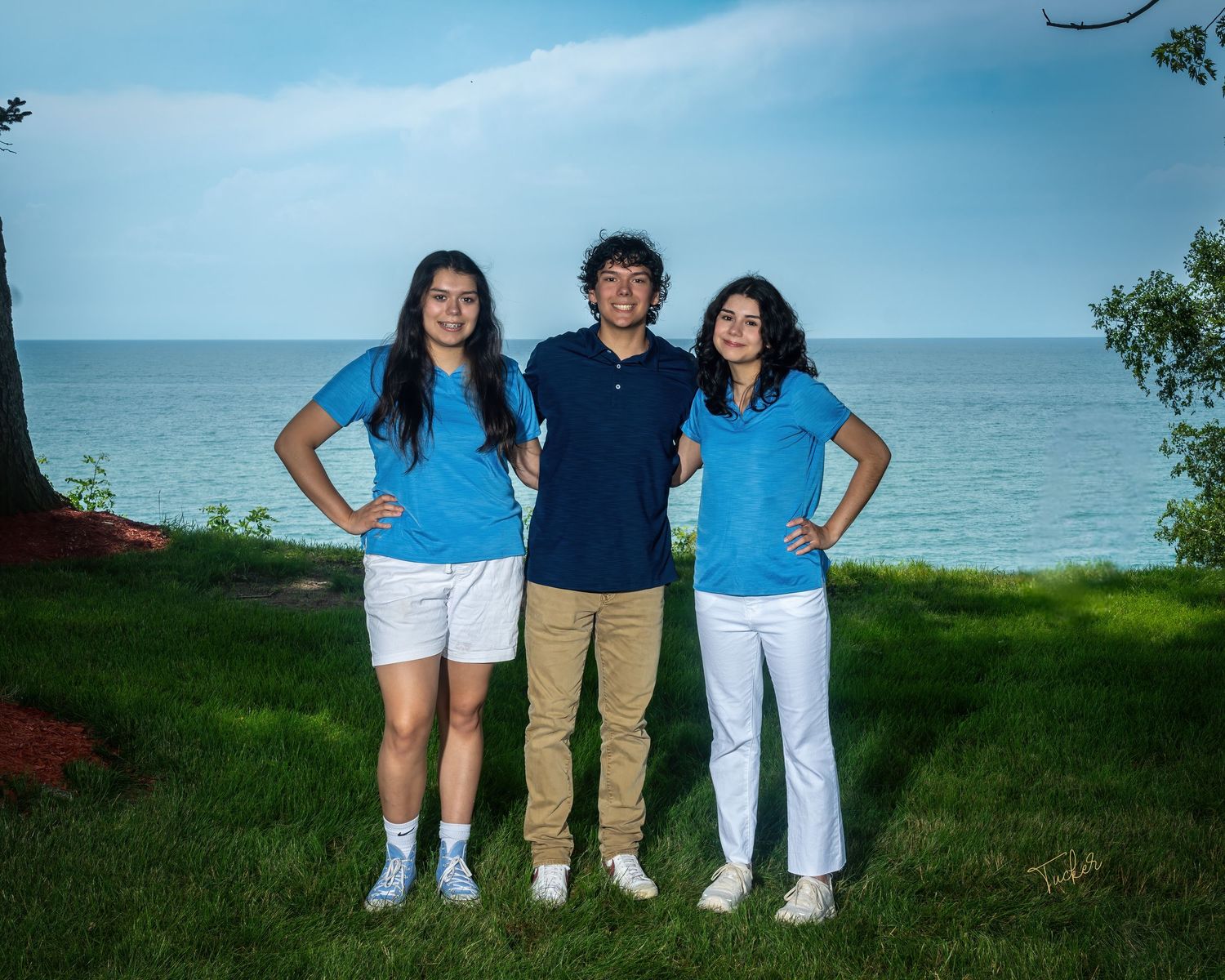 Three young adults standing on green grass with lake in background, wearing blue and white outfits.