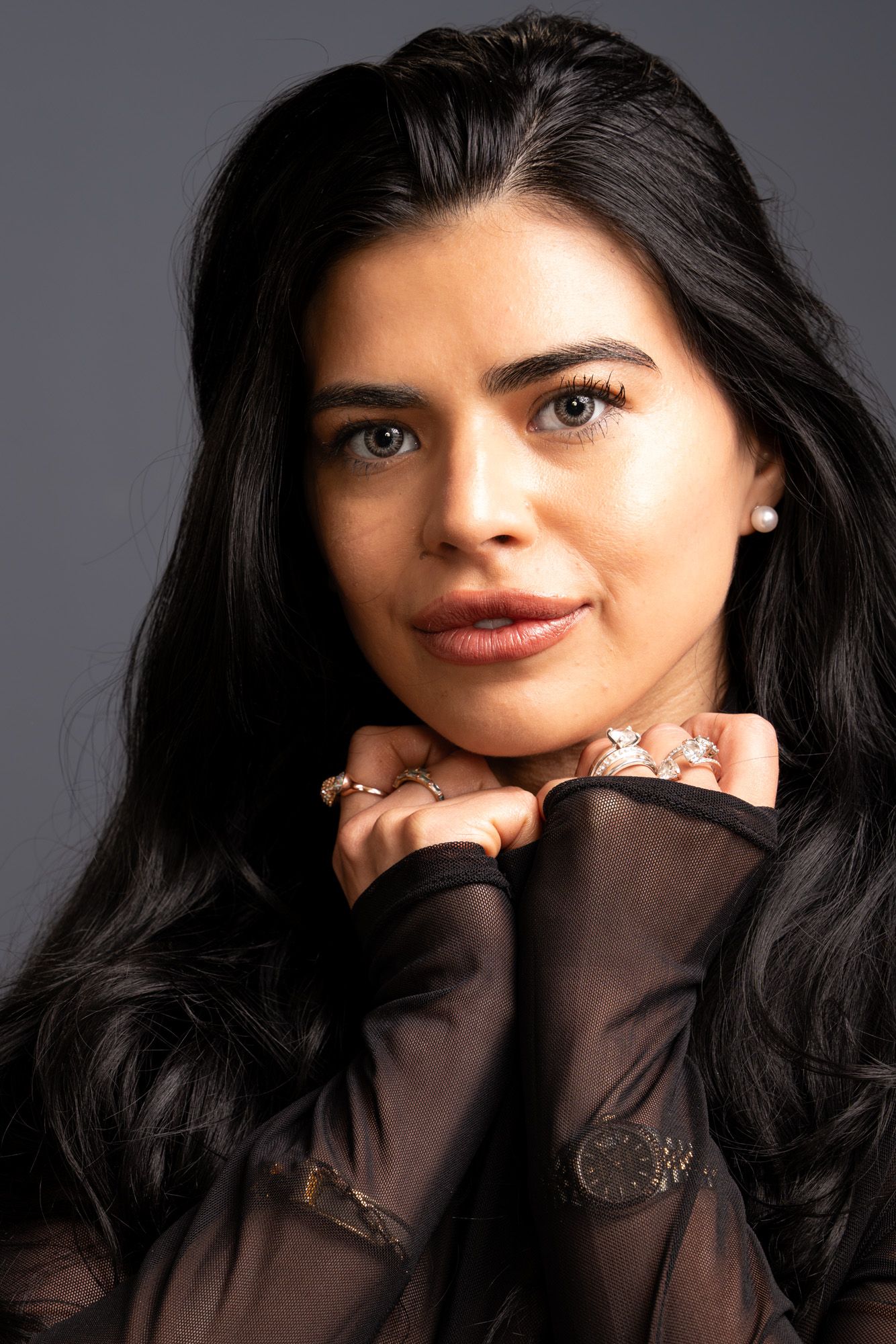 Elegant young woman with long black hair and rings posing with hands clasped near face.