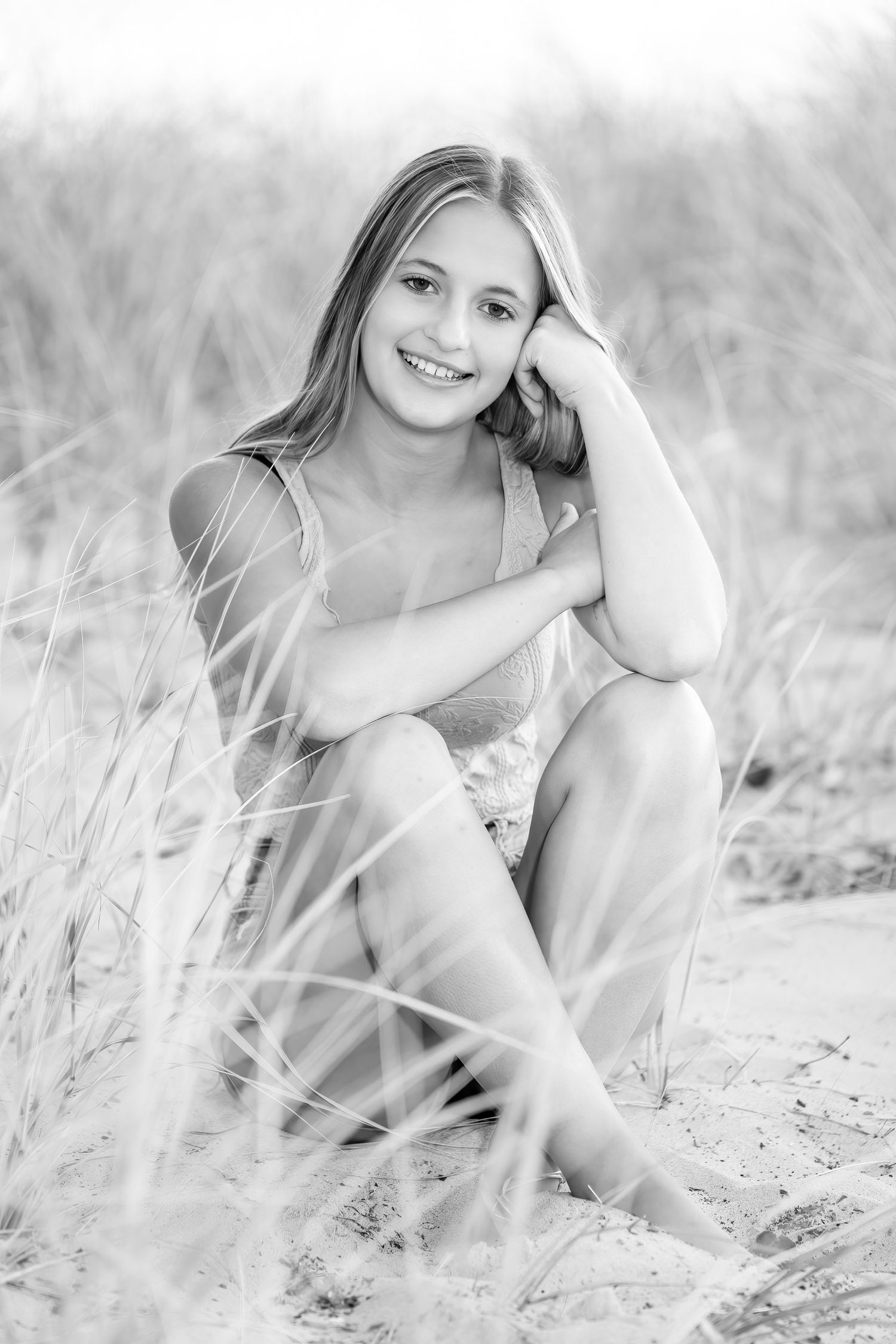 Black and white portrait of smiling teen girl sitting on sandy beach dunes with tall grass.