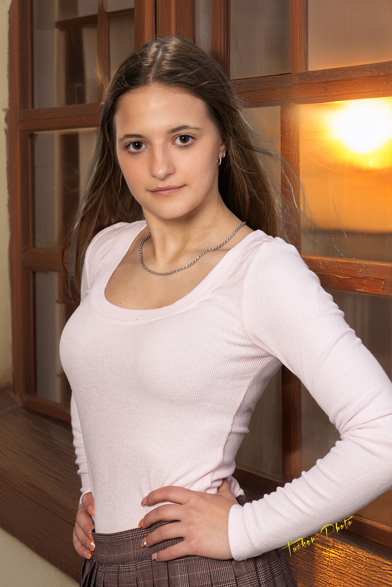 Young brunette woman in pink long-sleeve top and plaid skirt posing near window at sunset.