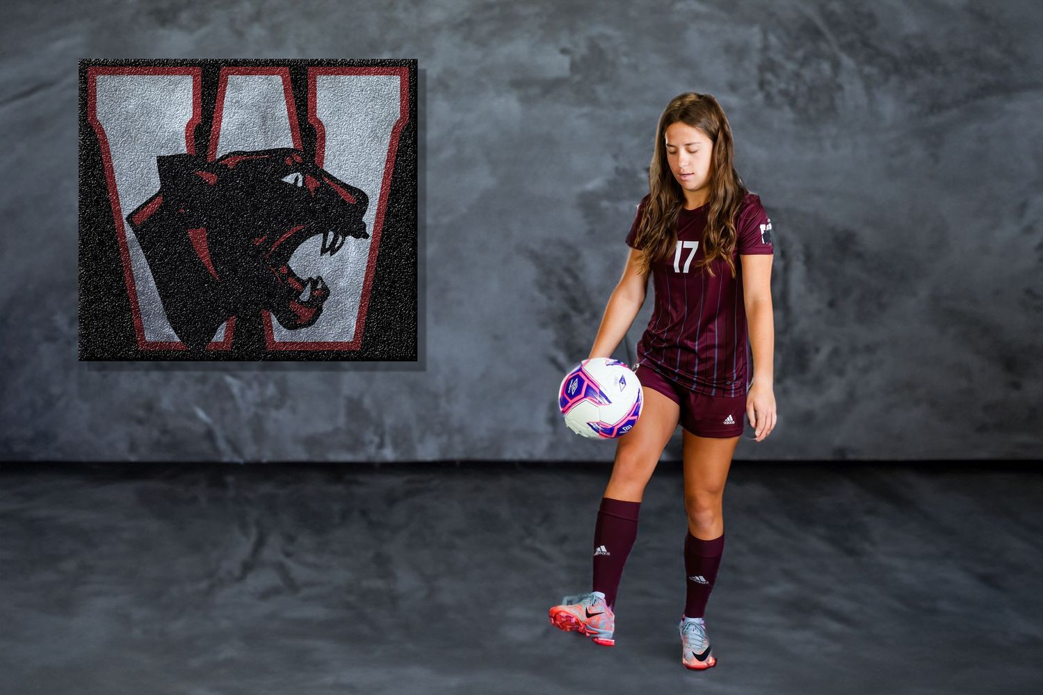 Female soccer player wearing number 17 jersey poses with ball in front of school mascot banner.