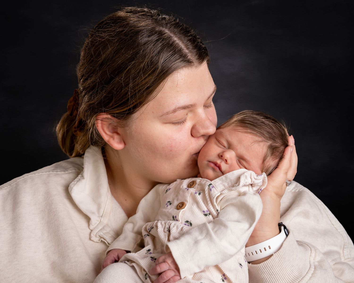 New mother gently kissing her sleeping newborn baby in a tender studio portrait.