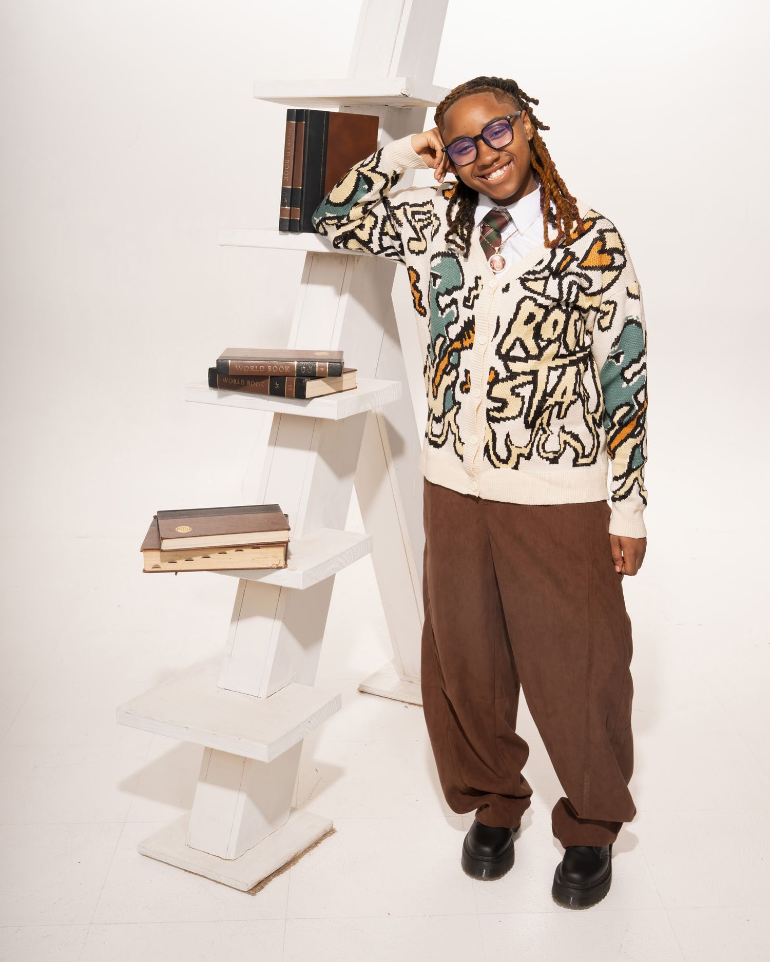 Smiling student posing beside white geometric bookshelf wearing graphic cardigan and brown trousers.