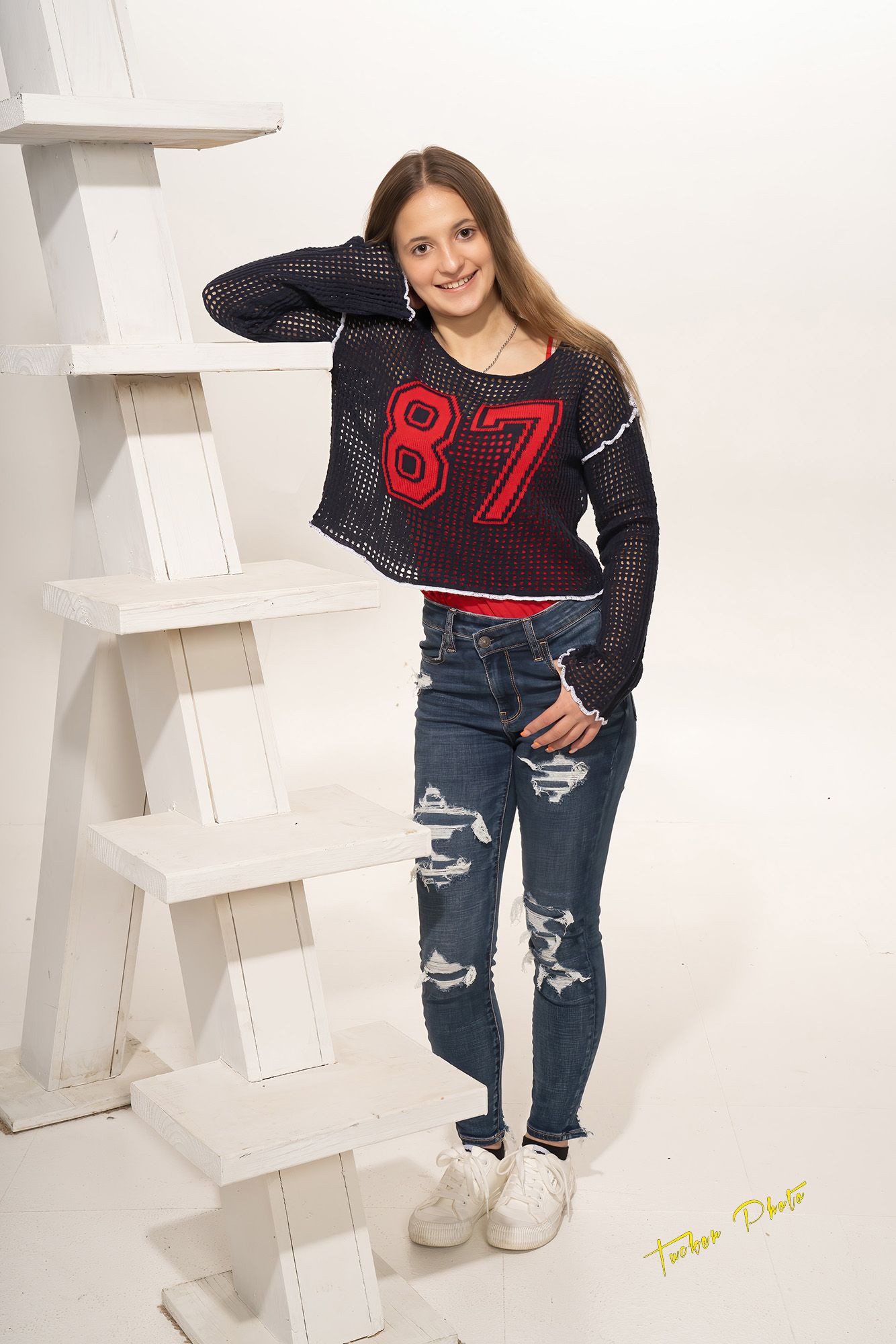 Teen girl in black mesh 87 jersey top and ripped jeans poses by white ladder in studio.
