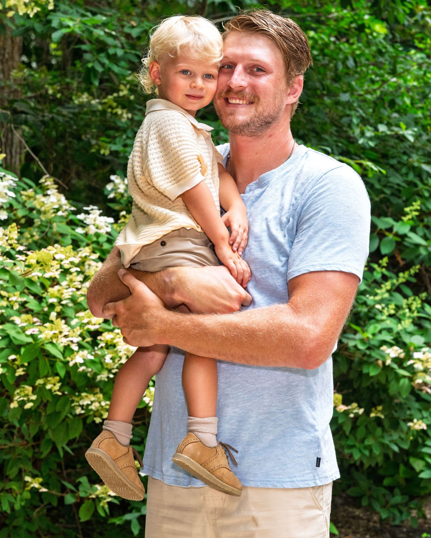 A smiling father holds his young blonde toddler son outdoors against a backdrop of green bushes.