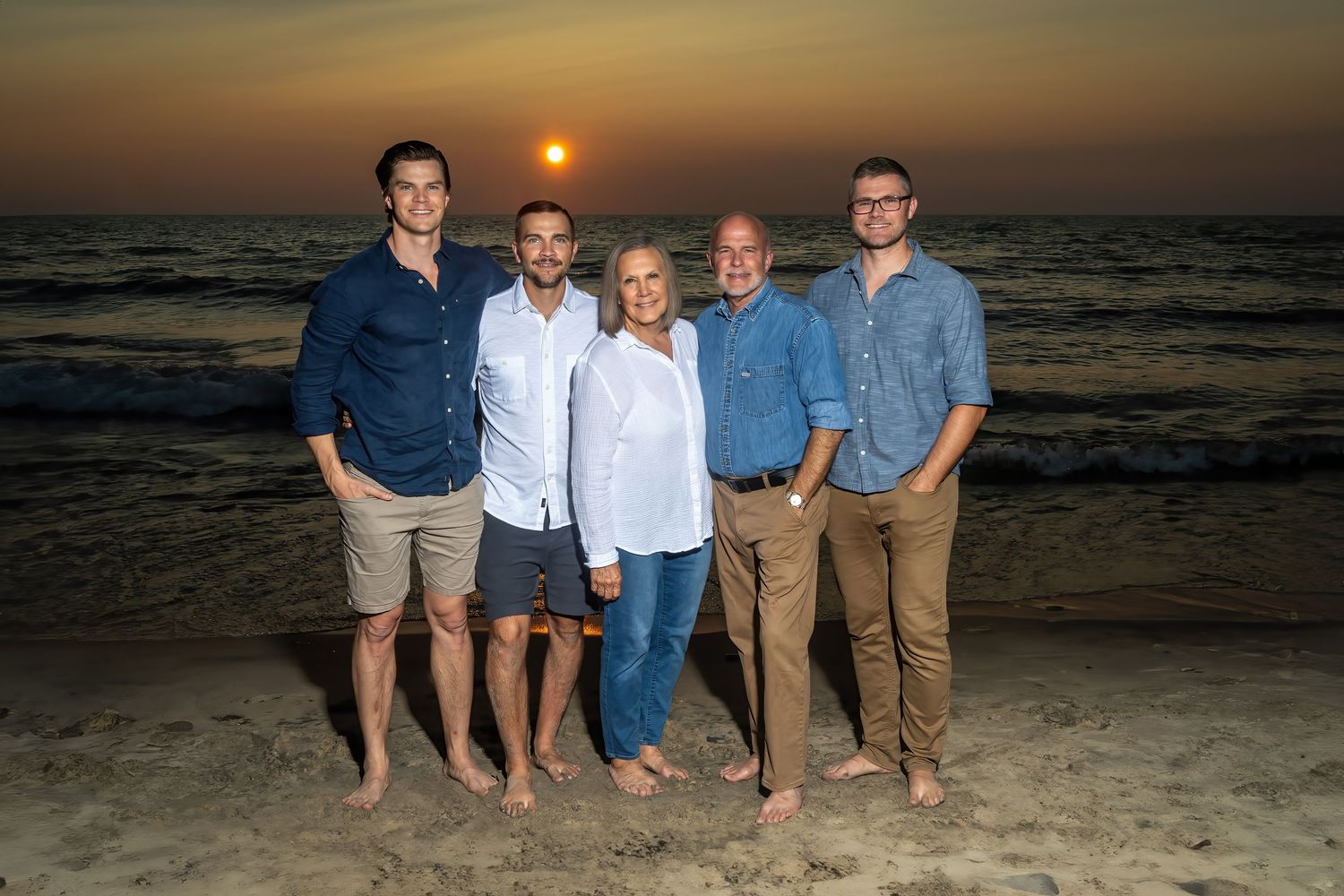 Family of five posing on beach at sunset with ocean waves in background.