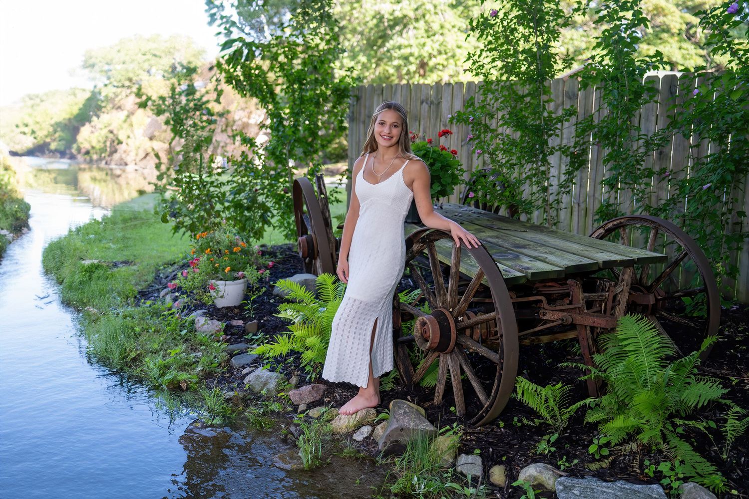 Blonde woman in elegant white maxi dress standing beside antique wagon wheel by a peaceful canal.
