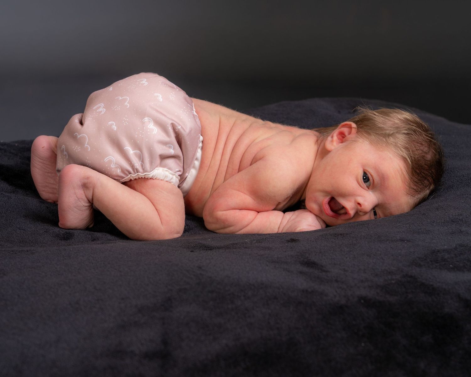 Newborn baby lying on dark blanket wearing diaper in professional studio photography.