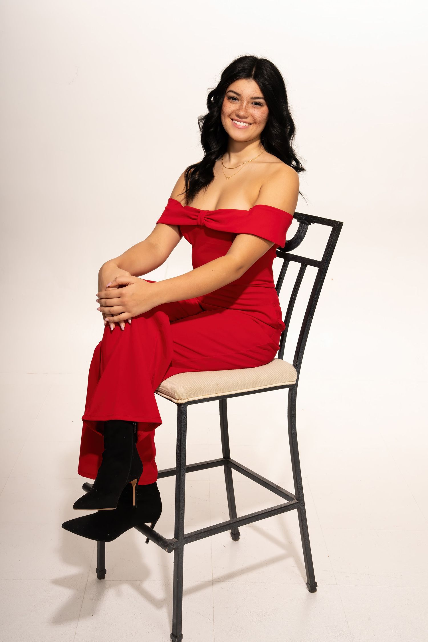 Smiling young woman in a red off-shoulder formal jumpsuit sitting on a bar chair in a white studio setting.