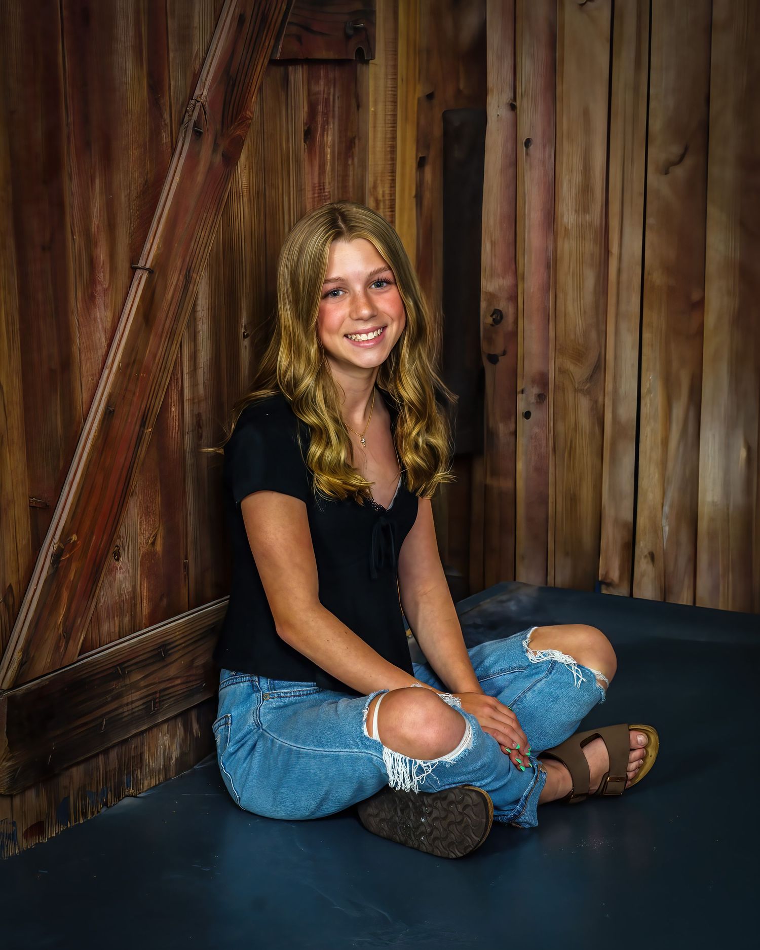 Smiling teenage girl in black top and ripped jeans sitting cross-legged on floor near rustic wooden barn wall.