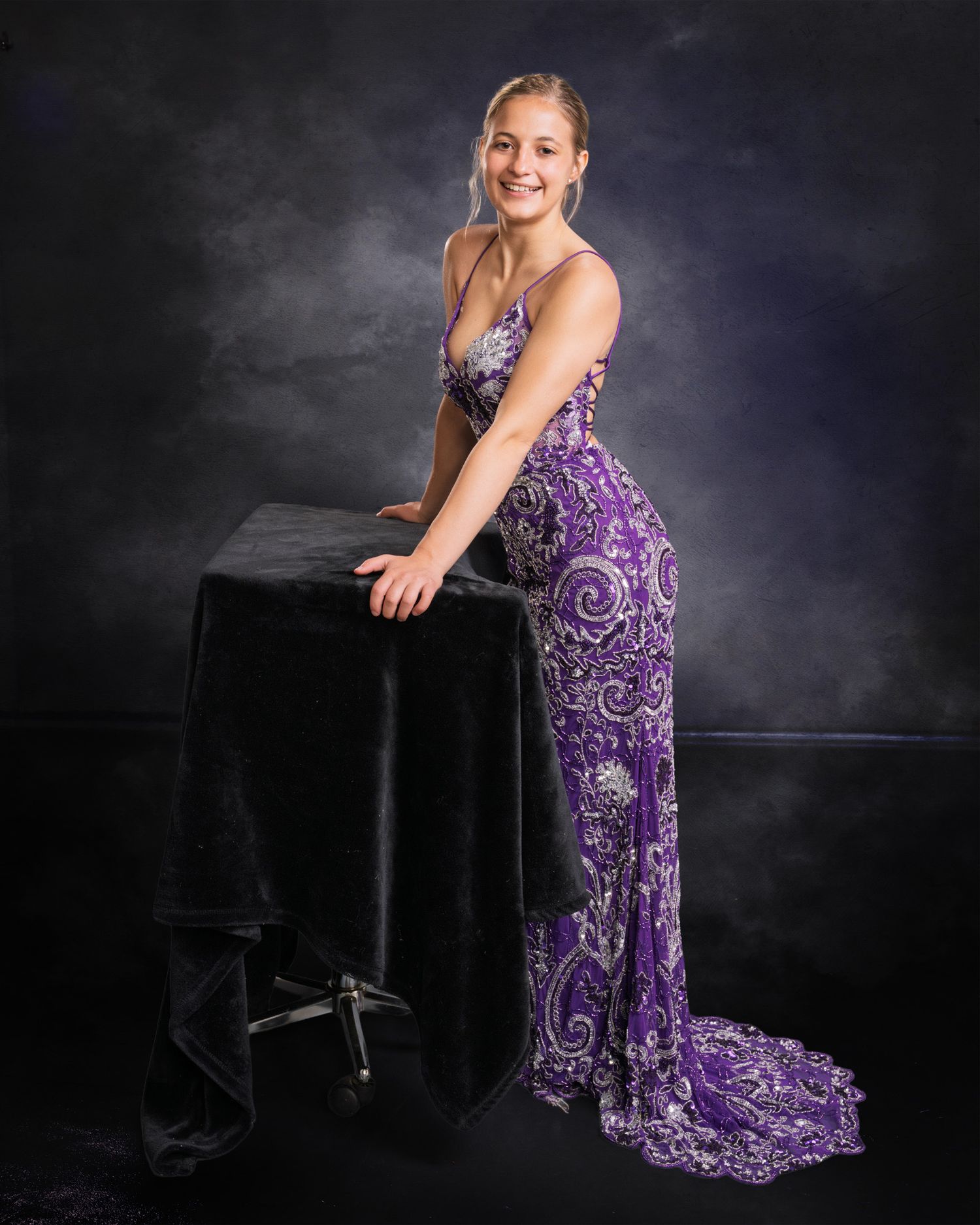 Smiling blonde teen in elegant purple beaded gown posing beside black velvet table for prom portrait.
