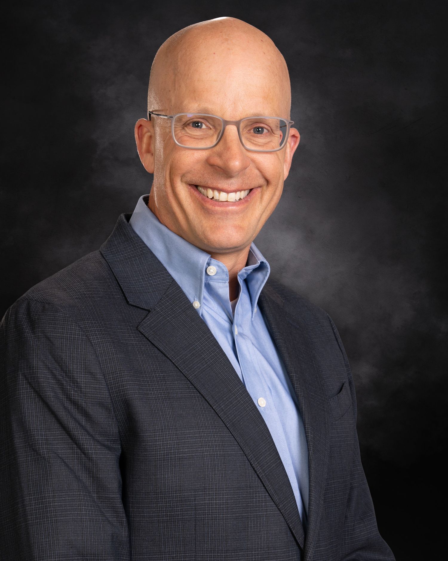 Professional portrait of a smiling bald man with glasses wearing a navy sport coat and light blue shirt.