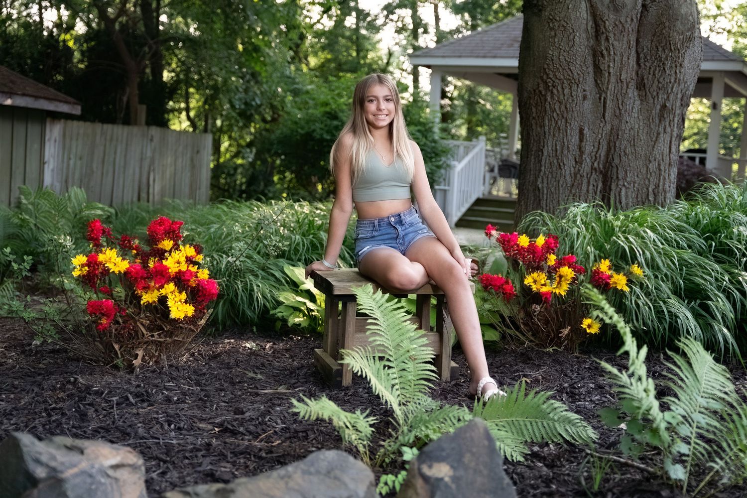 Blonde teen girl in crop top and denim shorts sits on garden bench surrounded by colorful flowers.