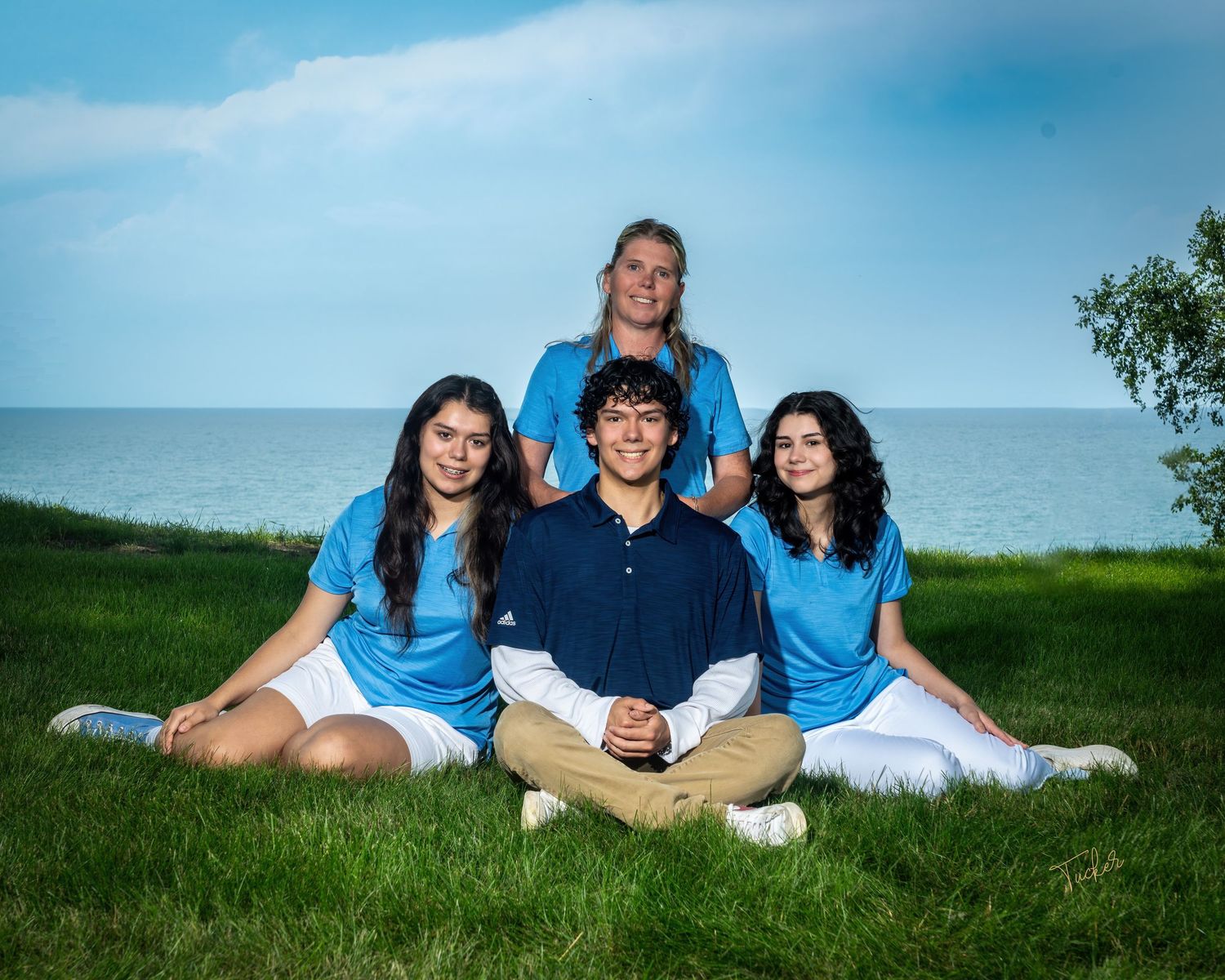 Four young adults posing on grass near lake, one standing behind three seated friends in blue shirts.