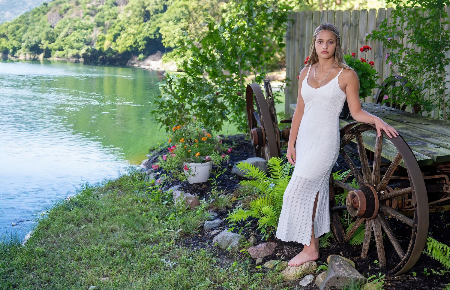 Young woman in white maxi dress poses beside rustic wagon wheel near scenic riverside garden setting.