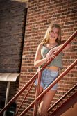 Blonde teenage girl in casual outfit poses on red metal staircase against brick building exterior.
