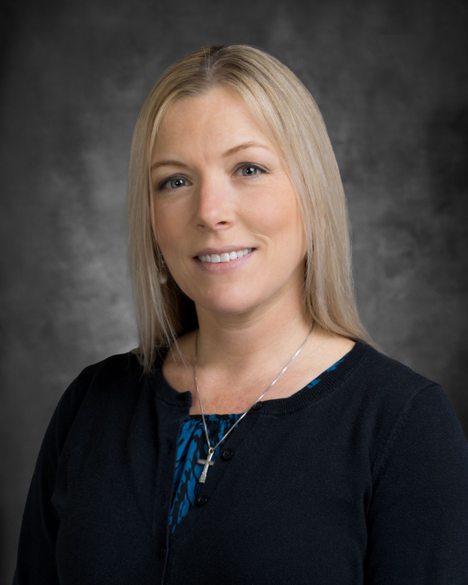 Professional headshot of a blonde woman wearing a black cardigan and cross necklace against a gray background.