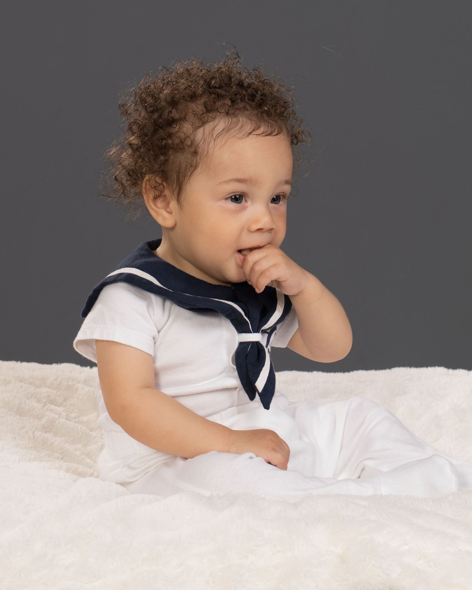 Adorable baby in white sailor outfit with navy collar sitting on fluffy white blanket.