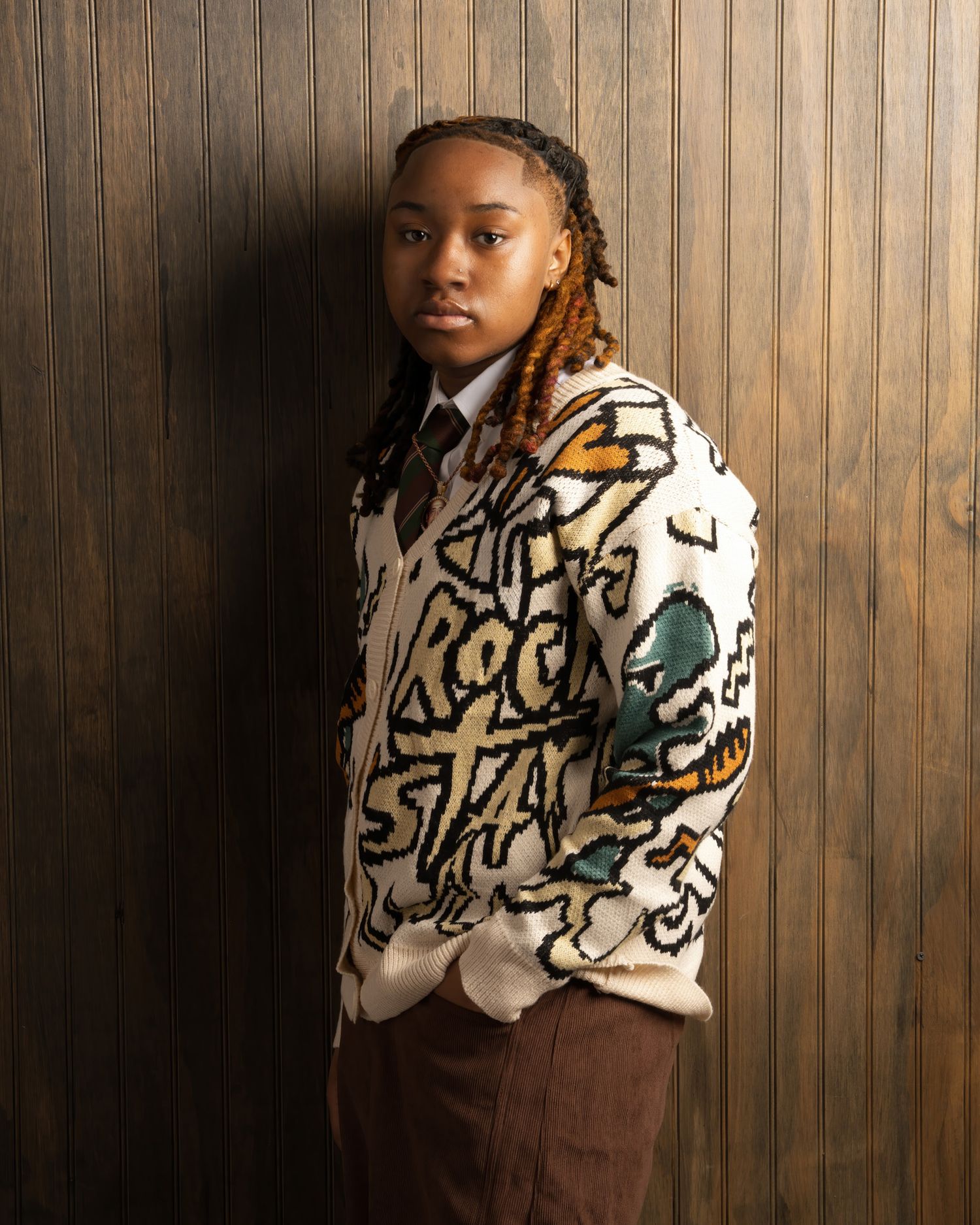 Young person in patterned graffiti cardigan with braids leaning against wood wall.