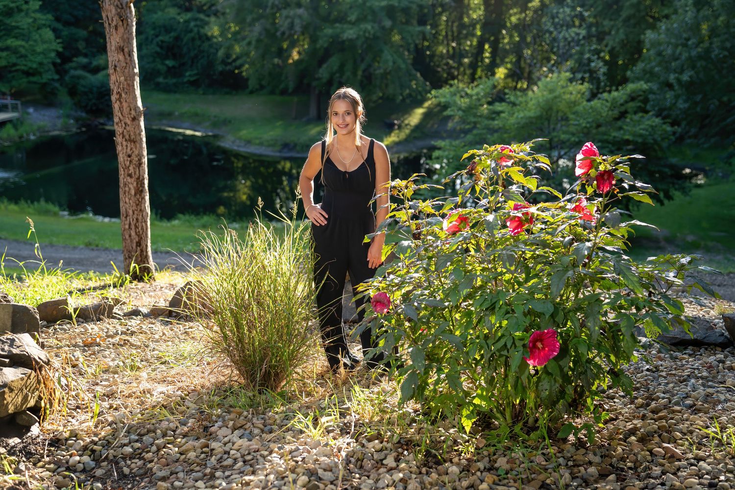 Young woman in black jumpsuit walks through sunlit garden with colorful roses and pond backdrop.