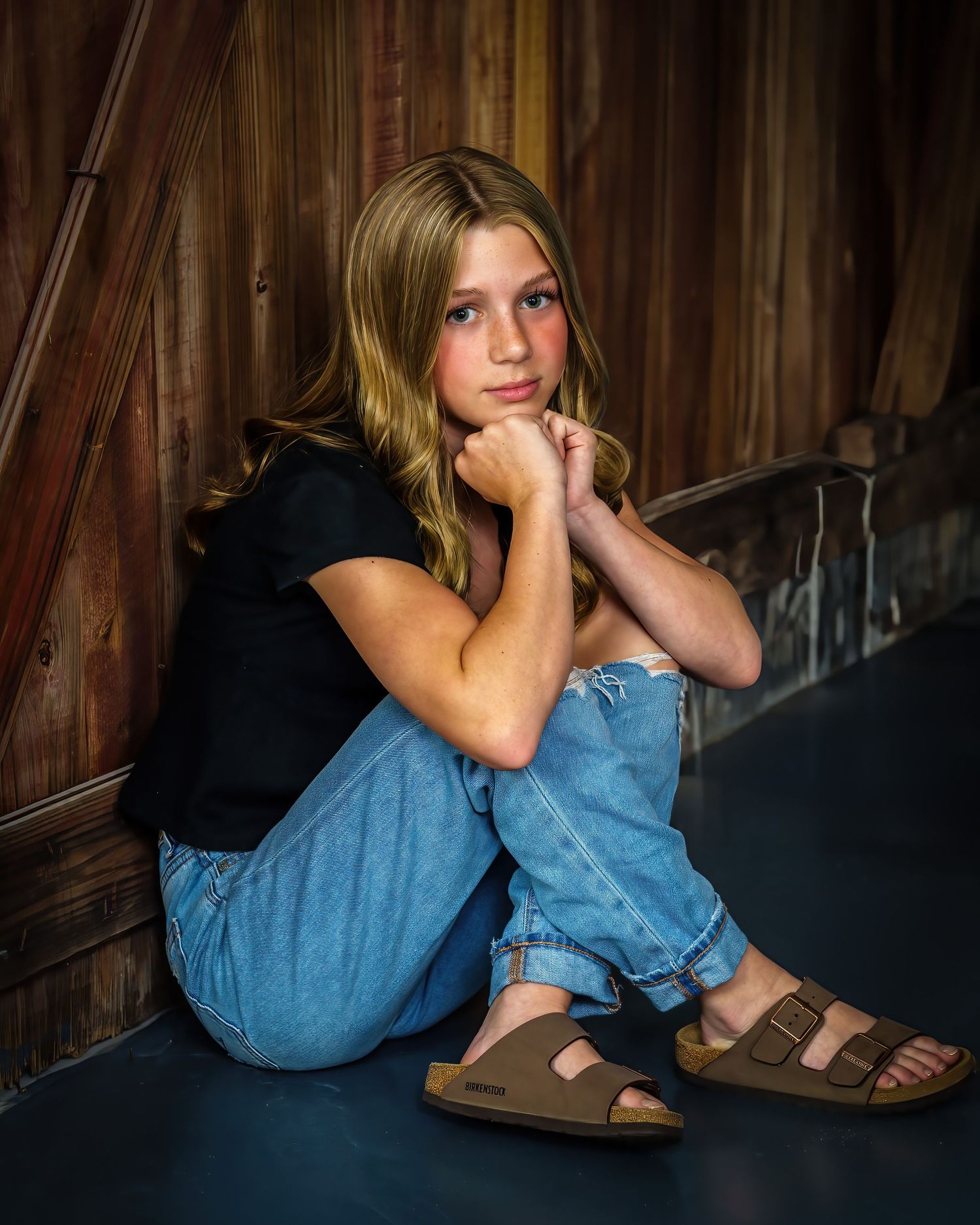 Teen girl in black top and jeans sitting pensively on floor against a warm rustic wooden barn backdrop.