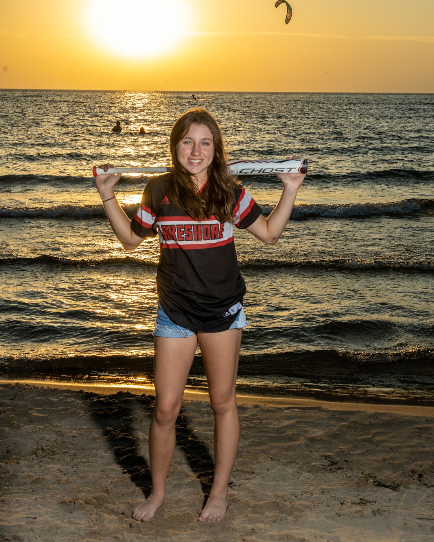 Teen girl in softball jersey holds bat at sunset beach.