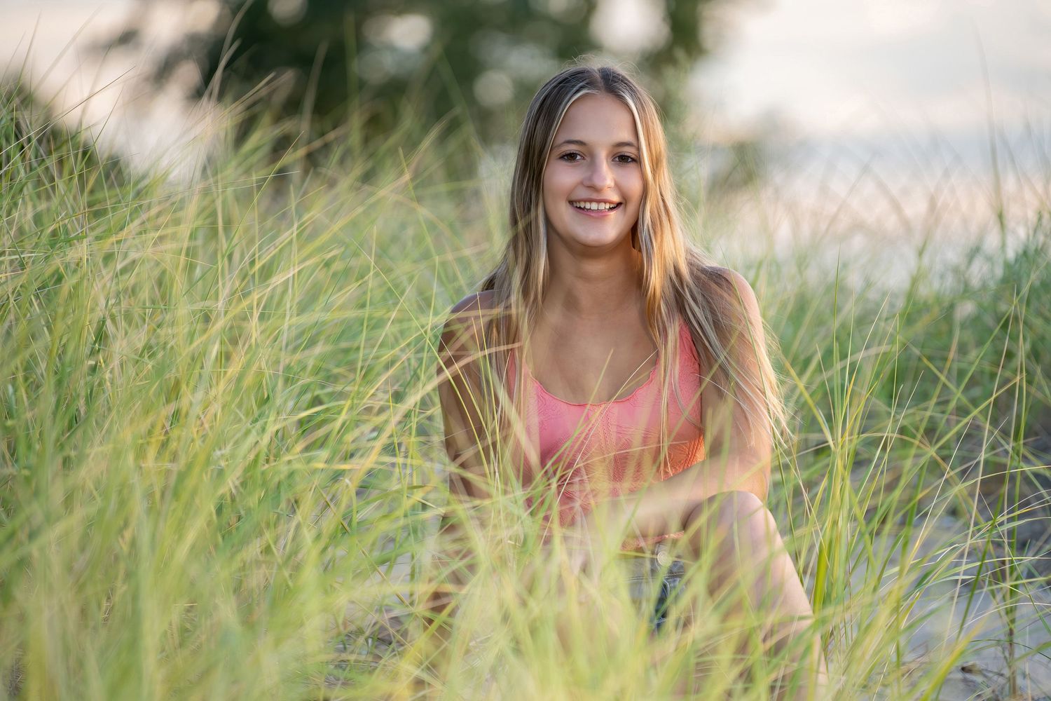Smiling young woman with long blonde hair sitting in tall beach grass at sunset.