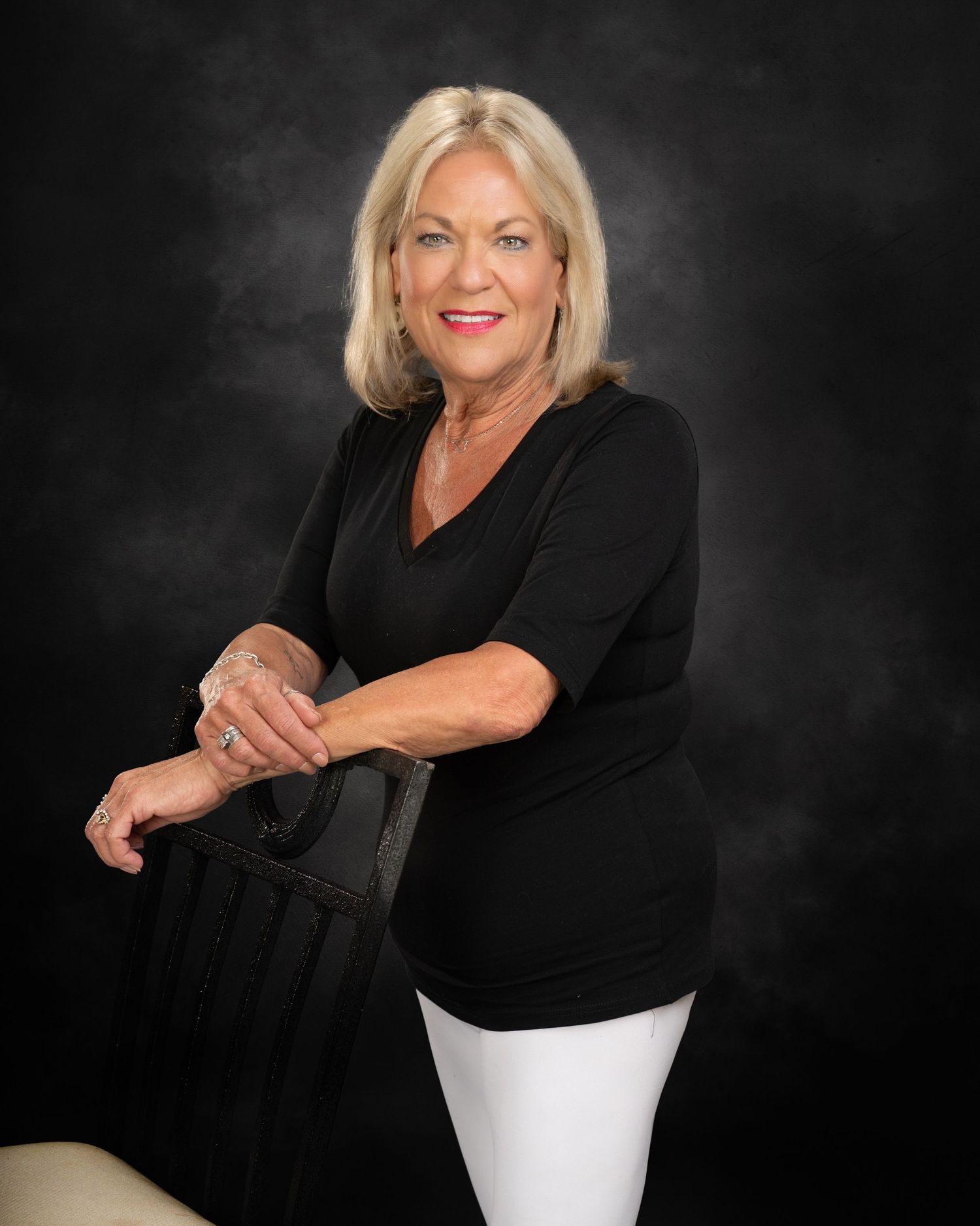 Confident blonde woman in black v-neck top and white pants posing beside chair on dark background.